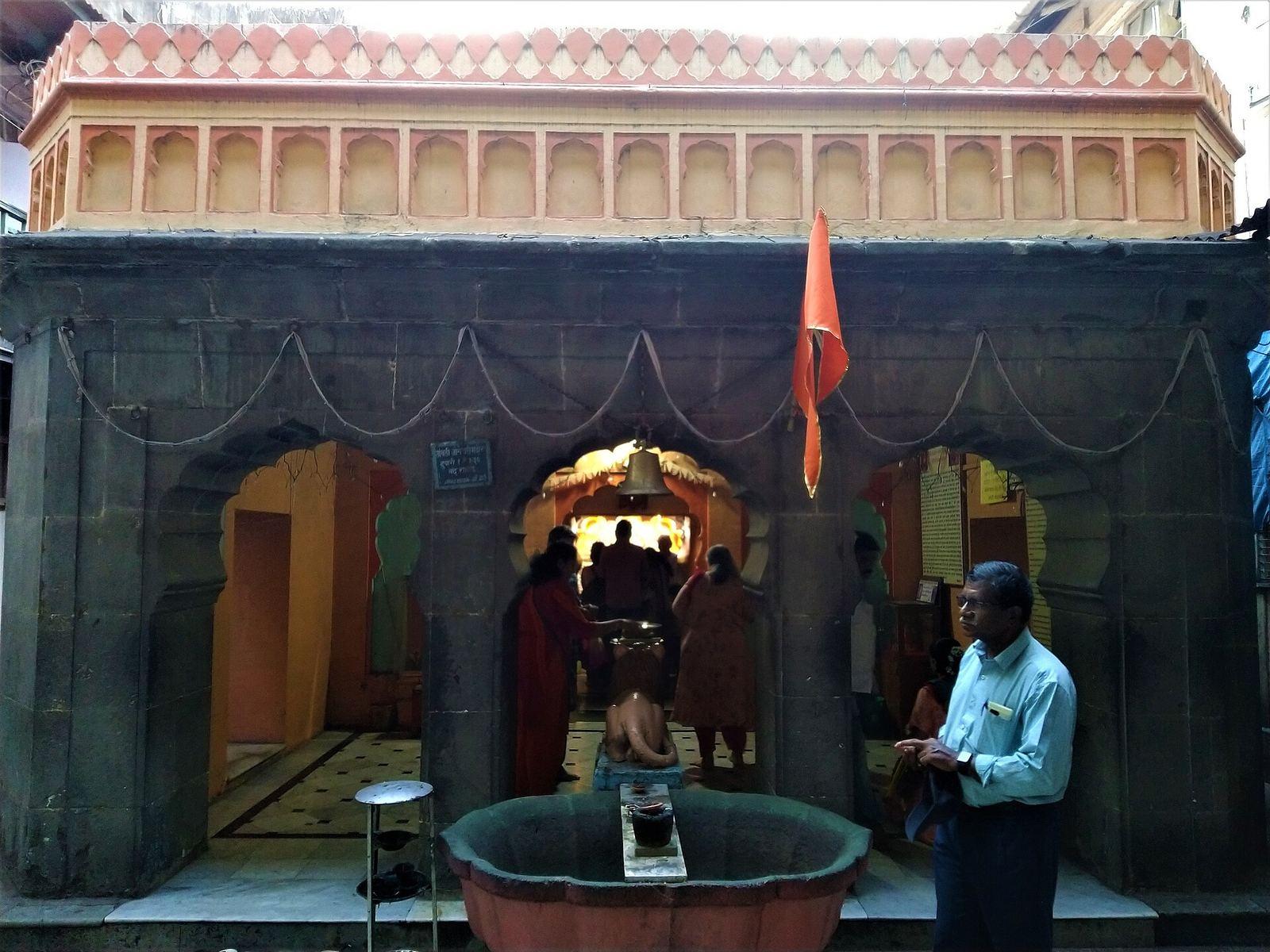 Street front of Tambdi Jogeshwari temple in Pune, with a carved stone arch, a lamp pillar, and orange flags